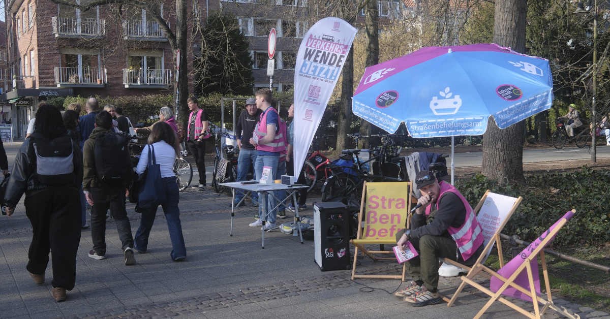 Eine Mahnwache der Initiative „Fahrradstadt.ms“ auf einem sonnigen, gepflasterten Platz in Münster. Im Vordergrund sitzt ein Aktivist mit rosa Warnweste und Mikrofon in einem Liegestuhl unter einem blau-pinken Sonnenschirm des „Straßenbefreiungsamts“. Daneben steht eine weiße Beachflag mit der Aufschrift „VERKEHRSWENDE JETZT!“. Im Hintergrund sind Passanten, Fahrräder und kahle Bäume vor städtischen Wohngebäuden zu sehen.