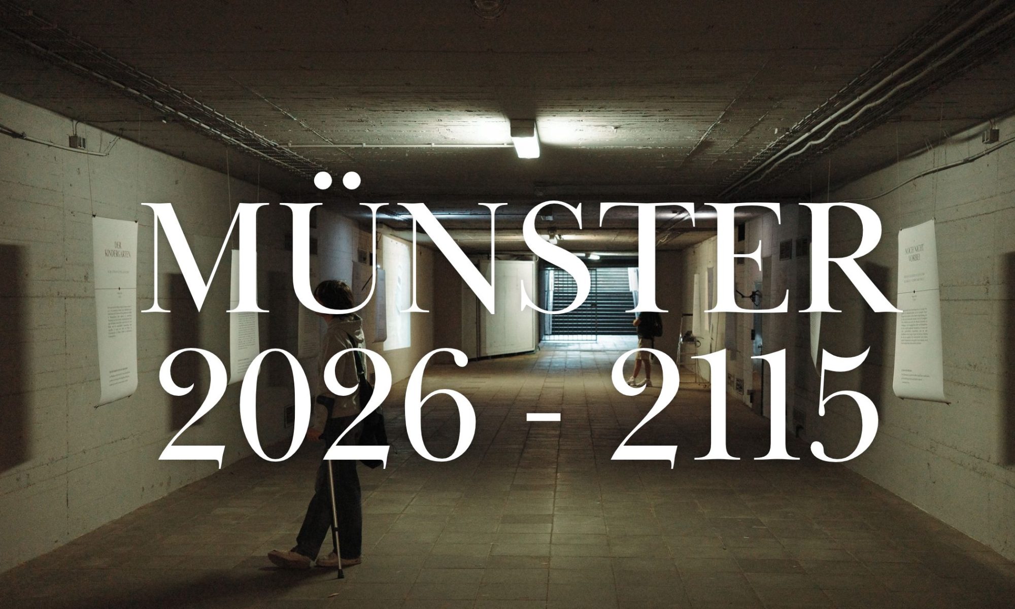 Ein Foto aus der Ausstellung Münster 2026-2115. Zu sehen sind mehrere Ausstellungsplakate in einem Tunnel mit nackten Betonwänden und -decken sowie Bodenfliesen. Die Stimmung ist kalt. Darüber der Schriftzug "Münster 2026-2115"